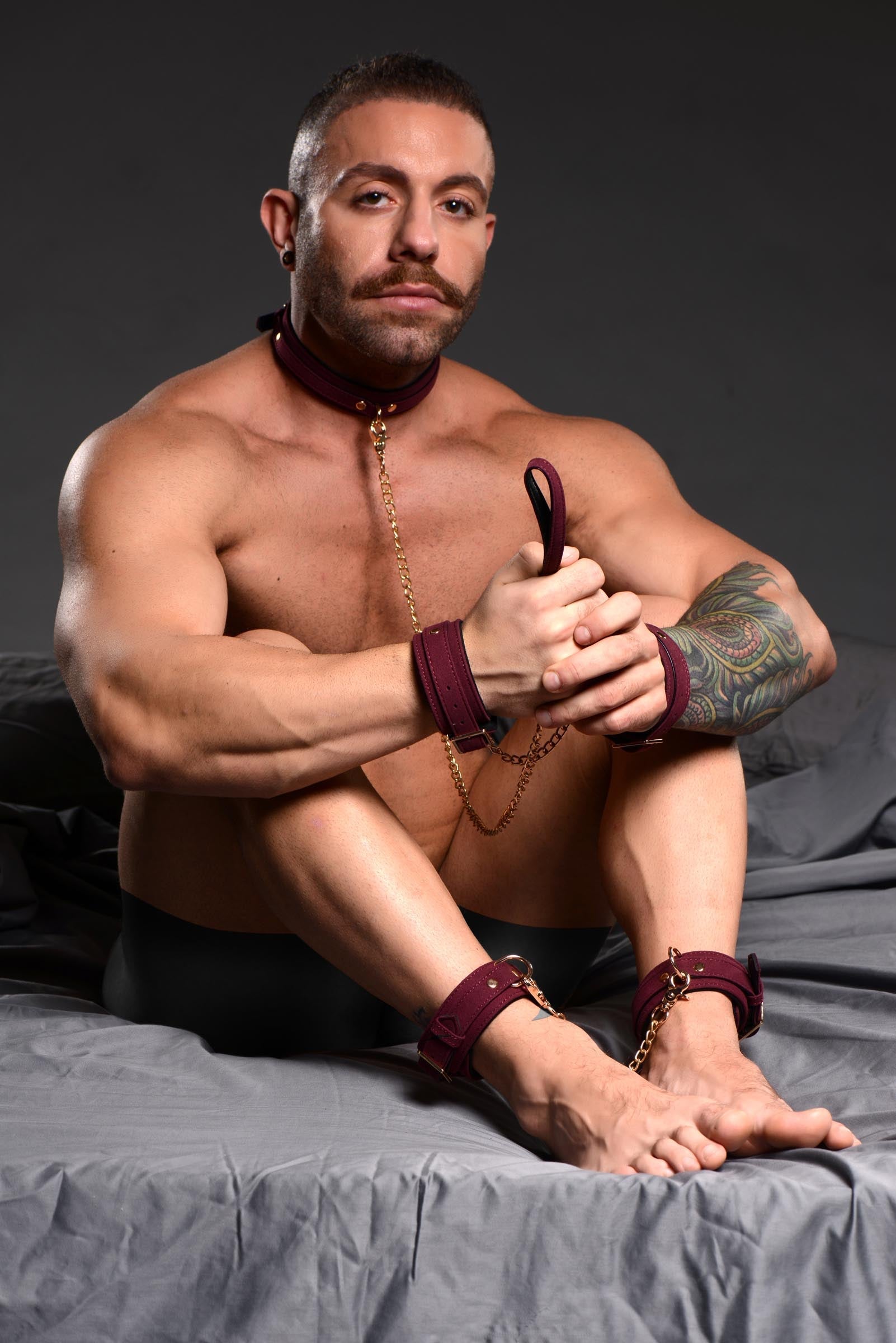 Shirtless male model on bed with wrists bound by velvet cuffs